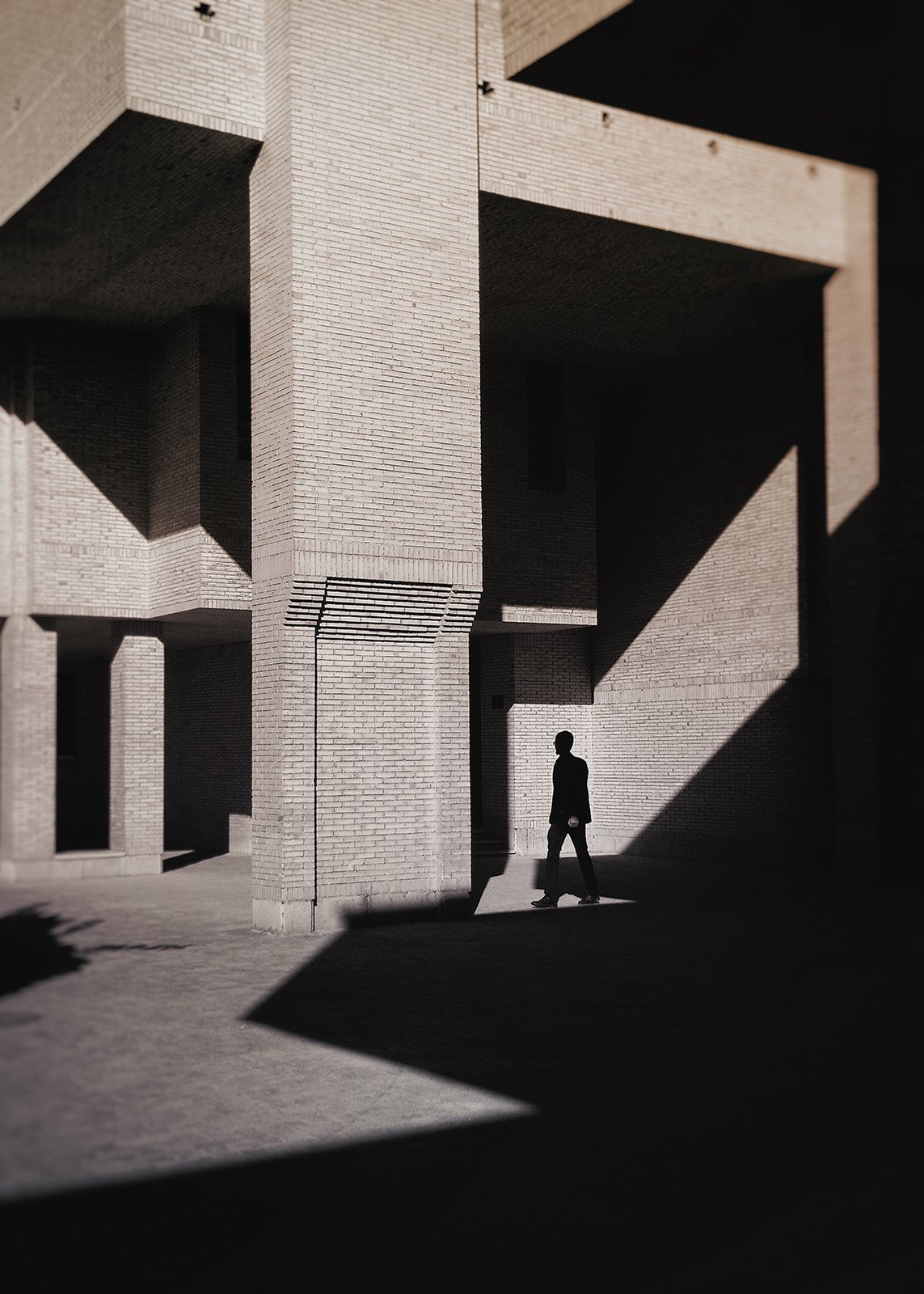 A man in silhouette walks through a sharp triangle of light between brutalist brick pillars in the 'Urban Eclipse' series.